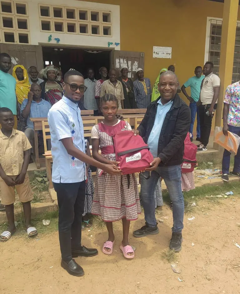 Remise d'un sac à dos scolaire SUSCOM à une élève devant l'école, en présence des villageois