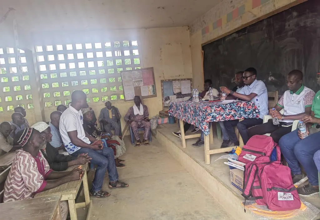 Réunion de village dans une salle de classe avec des sacs à dos SUSCOM posés devant l'estrade